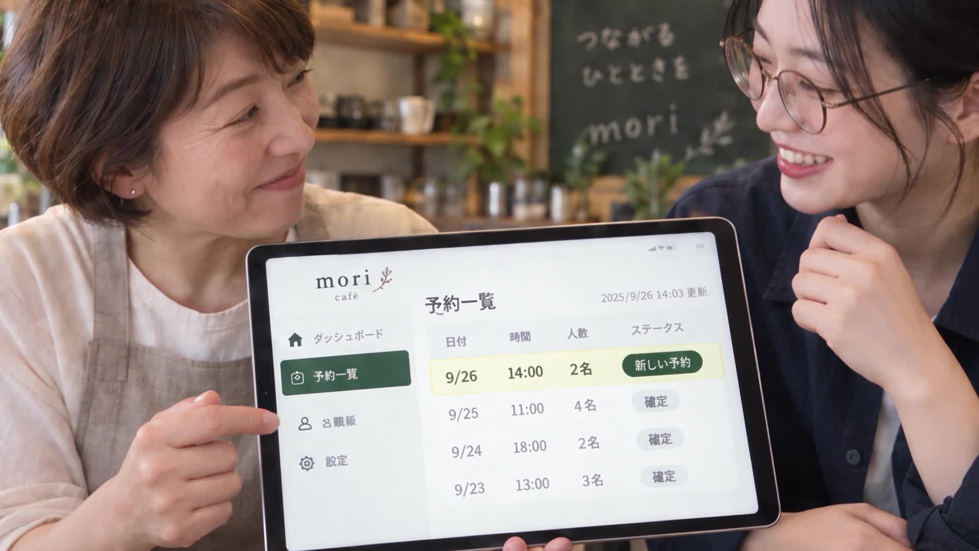 カフェの店主と学ぶ人が、タブレットの予約アプリを一緒に見て笑っている